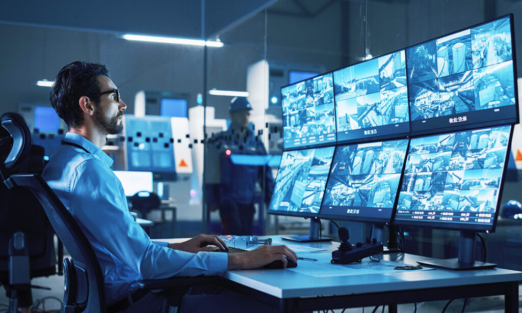 Industry 4.0 Modern Factory: Security Operator Controls Proper Functioning of Workshop Production Line, Uses Computer with Screens Showing Surveillance Camera Feed. High-Tech Security